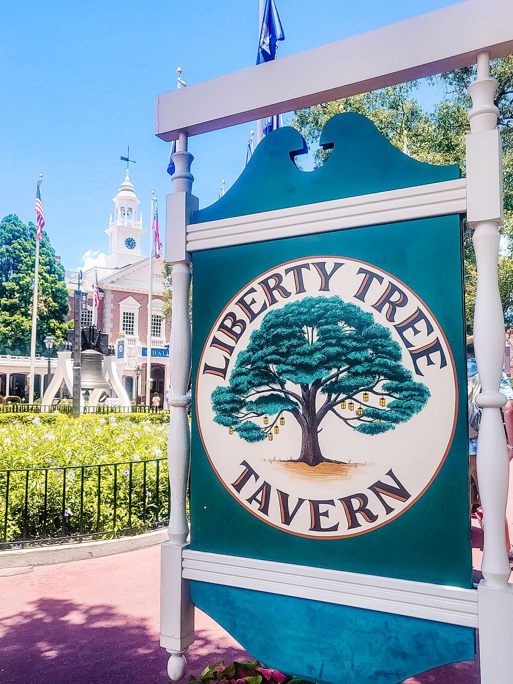Sign for Liberty Tree Tavern with colonial building and church steeple visible in background at Disney's Magic Kingdom.