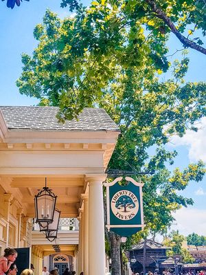 A charming storefront with a green Marketplace sign, hanging lantern, and leafy tree branches overhead on a sunny day.