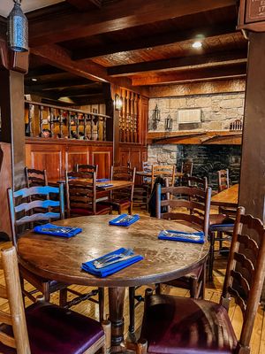 Rustic restaurant interior with wooden tables, ladder-back chairs, stone walls and blue placemats in a cozy dining room setting.