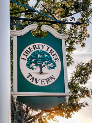 A vintage-style green and white hanging tavern sign for Liberty Tree Tavern featuring an oak tree logo at dusk.