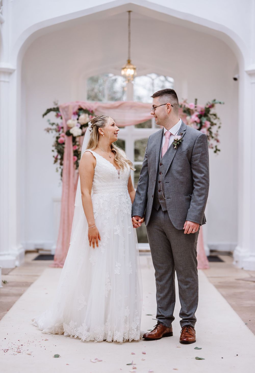 Bride and groom holding hands smiling at each other in front of a floral arch with pink draping at St Audries wedding venue.