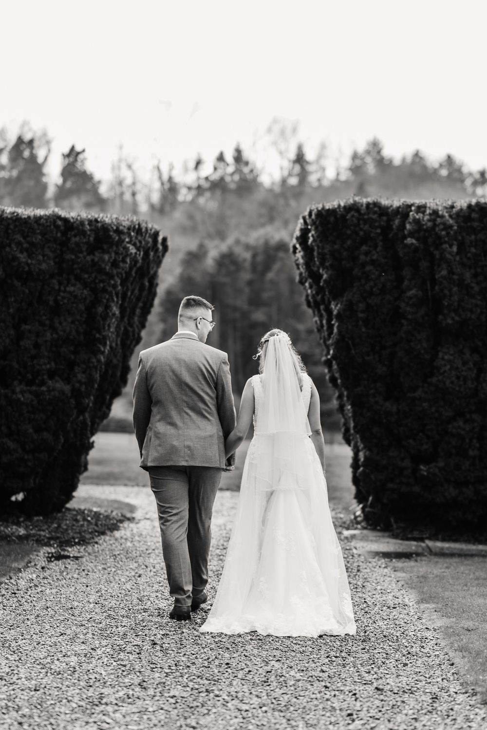 Black and white photo of a bride and groom walking hand in hand down a gravel path lined with tall hedges at St Audries Park, Wedding venue