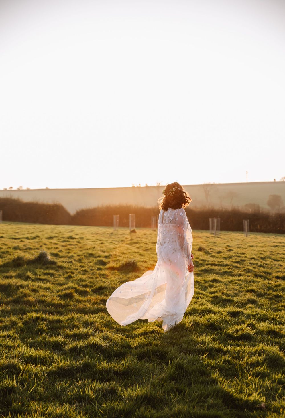 Bride in flowing lace wedding dress standing in sunlit green field at golden hour, dress billowing in the breeze at The Cornish Place.