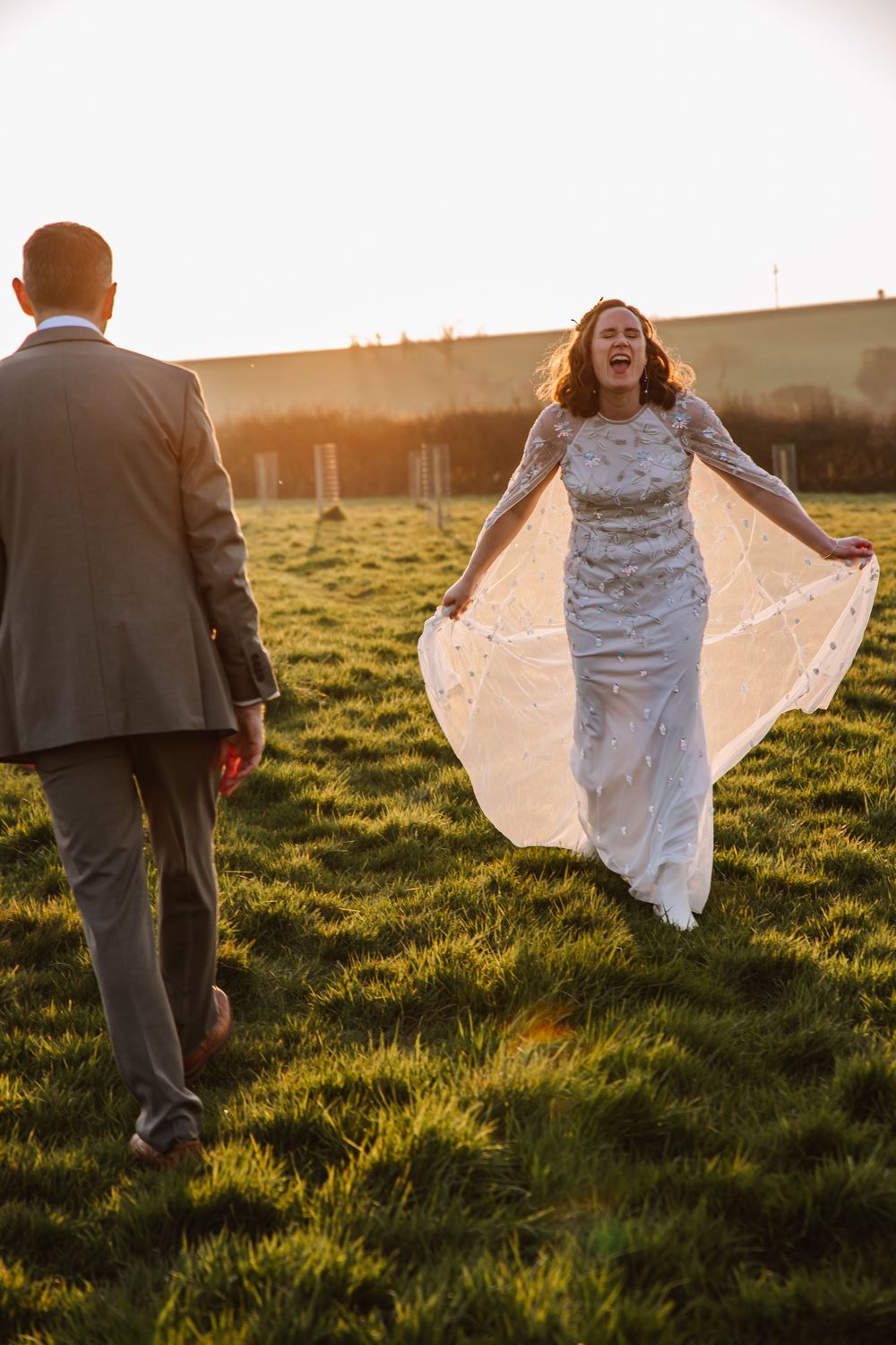Bride in lace gown holds dress while walking toward groom in grey suit during golden hour on green field at The Cornish Place, Wedding venue