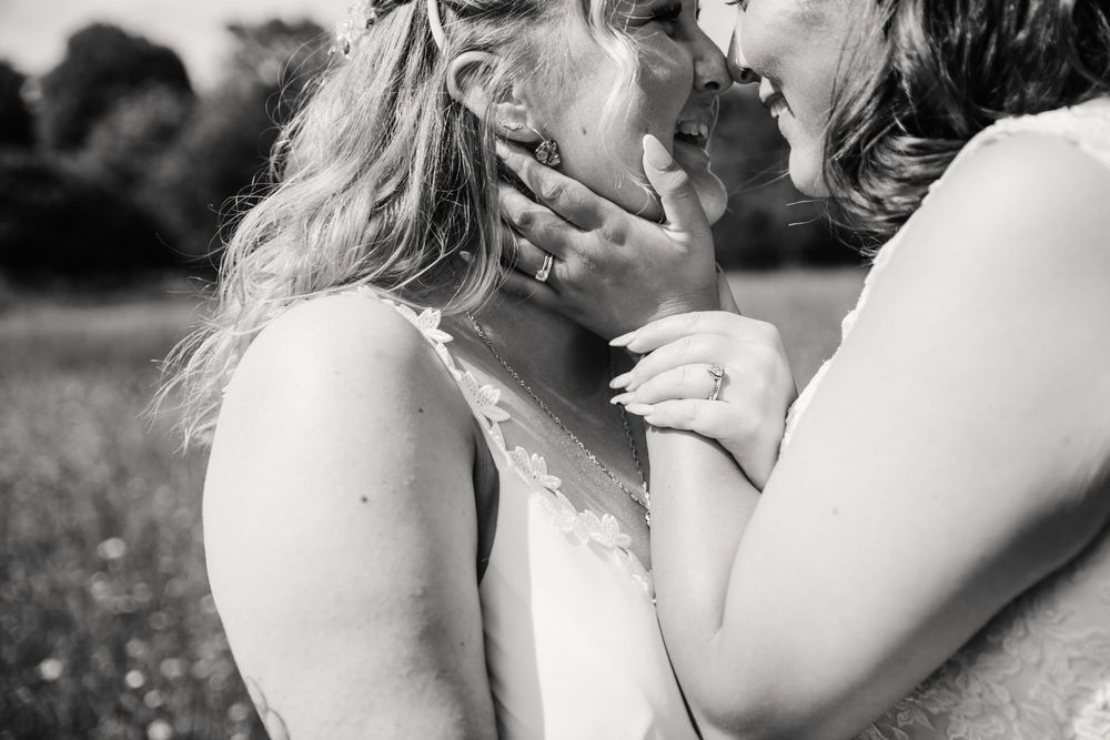 Two brides in white wedding dresses hold hands walking down a sunny road lined with green trees. LGBTQ+ tipi wedding at The Gaggle of Geese