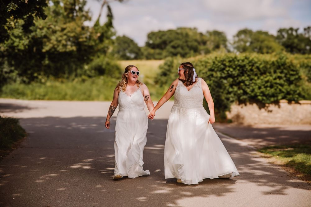 Two brides in white wedding dresses hold hands walking down a sunny road lined with green trees. LGBTQ+ tipi wedding at The Gaggle of Geese