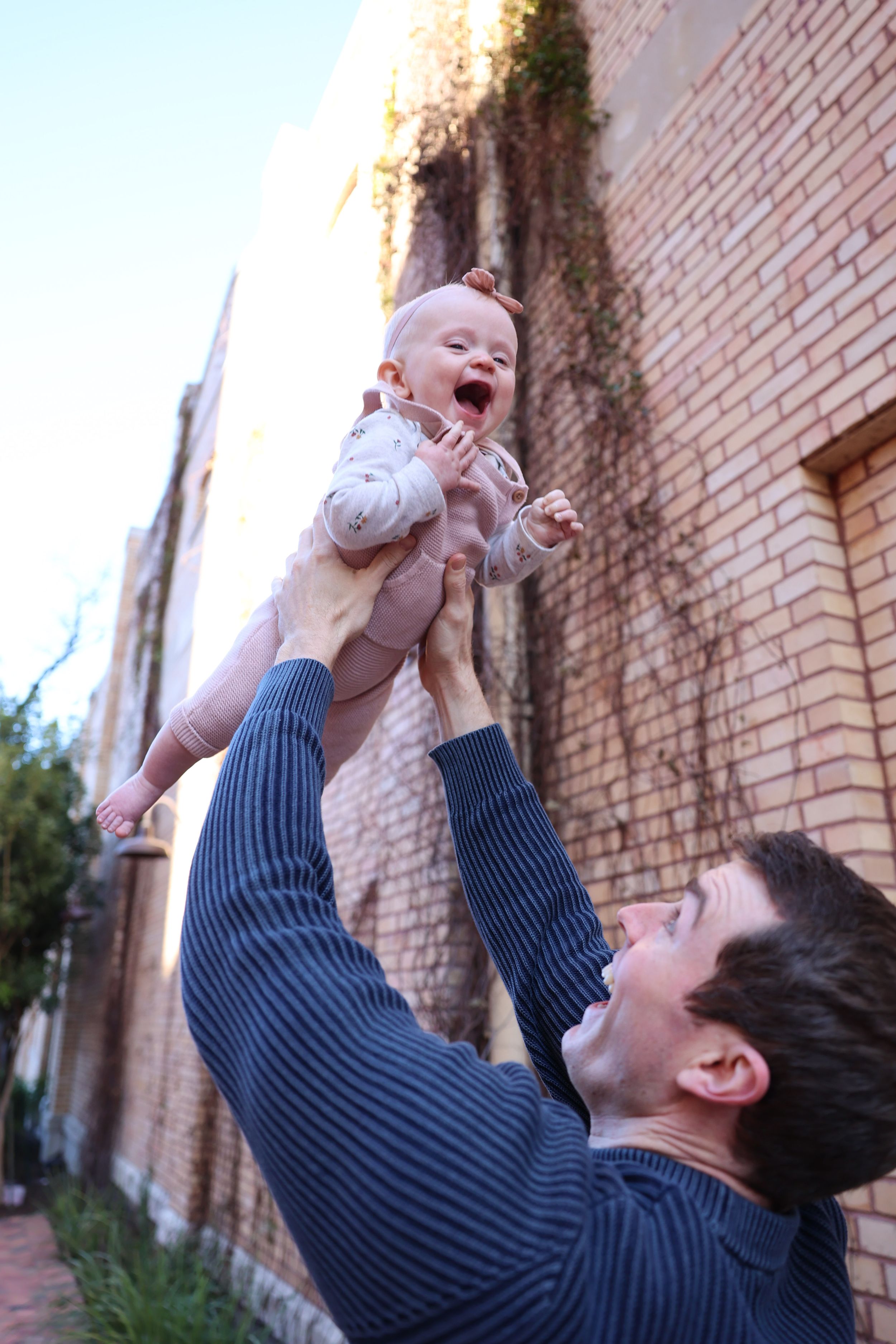 Baby laughing with joy while being lifted high into the air against a brick wall background.