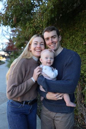A young couple in cozy sweaters holds their smiling baby outdoors near the River Walk in San Antonio, TX.