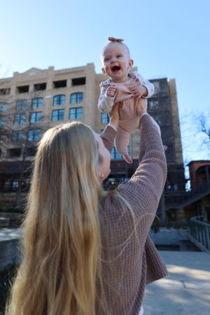 A person with long blonde hair lifts a joyful baby wearing beige clothing against a building exterior on a sunny day.