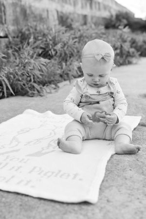 Sweet baby in overalls and bow sits on a blanket outdoors while looking down in this black and white portrait.