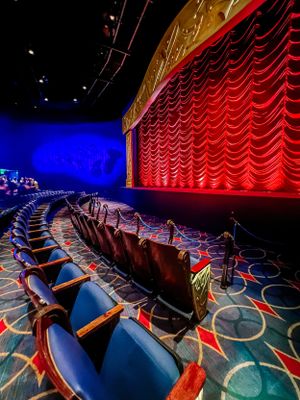 Empty rows of plush blue theater seats face a red curtained stage with elegant lighting in an entertainment venue.