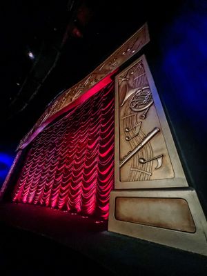 Red velvet curtain and ornate decorative panel illuminated with dramatic lighting in a theater or performance venue.
