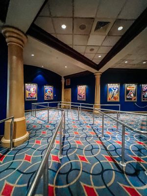 Theme park queue area with patterned carpet, decorative columns and movie posters displayed along blue-lit walls.