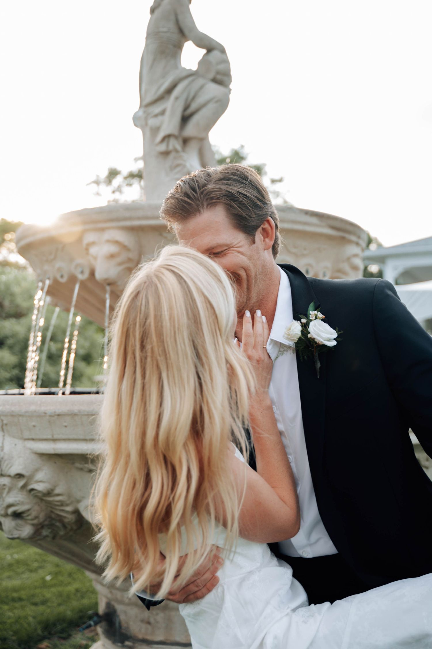 A couple shares a tender moment in their wedding attire on a lush green lawn with tall trees in the background.