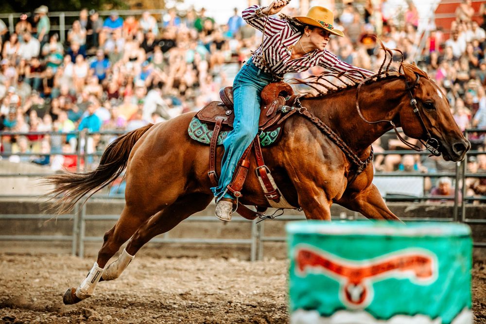Rodeo competitor in turquoise and brown rides a galloping brown horse around a barrel in an arena with spectators.