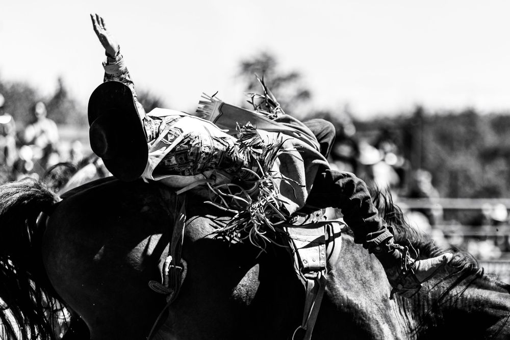 A dramatic black and white action shot of a rodeo rider on a bucking horse at a competitive event.