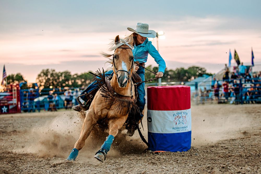 Barrel racing competitor navigates around blue and red barrels in a dusty rodeo arena during competition.