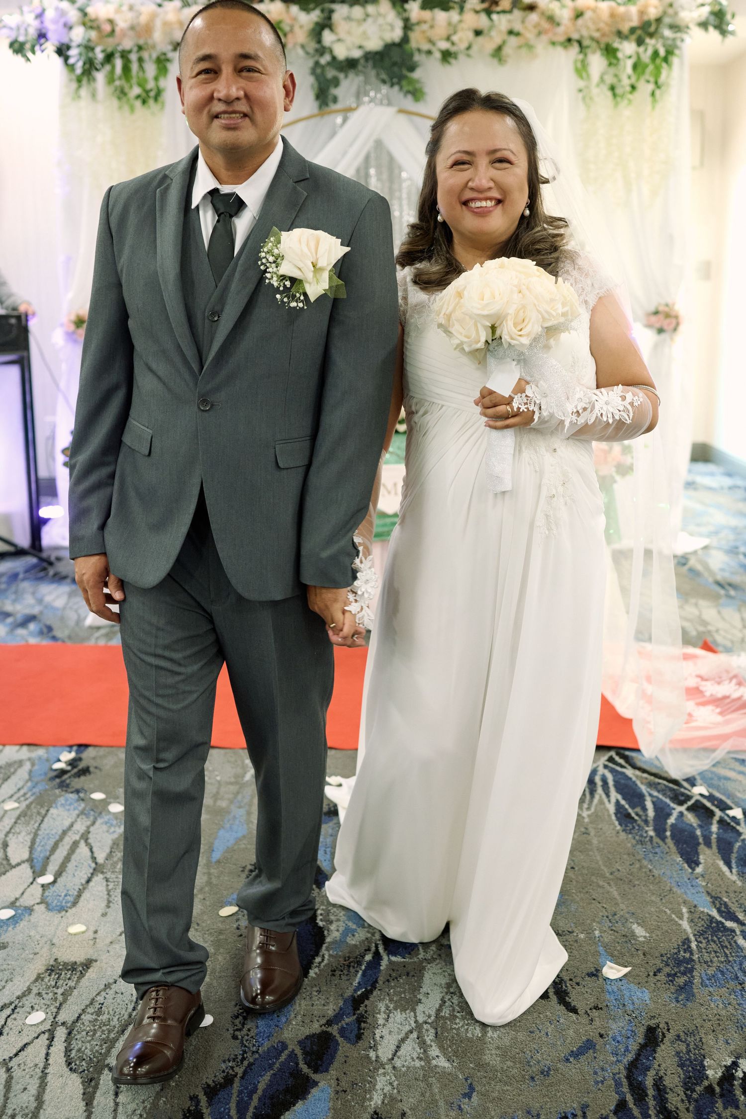 A wedding couple stands together at their ceremony, with the bride in a white gown and groom in a gray suit.