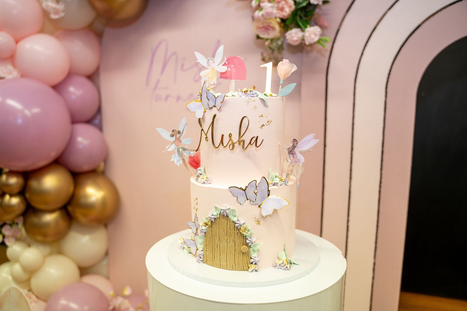 A whimsical pink and gold birthday cake decorated with delicate butterflies and floral accents sits on a white pedestal.