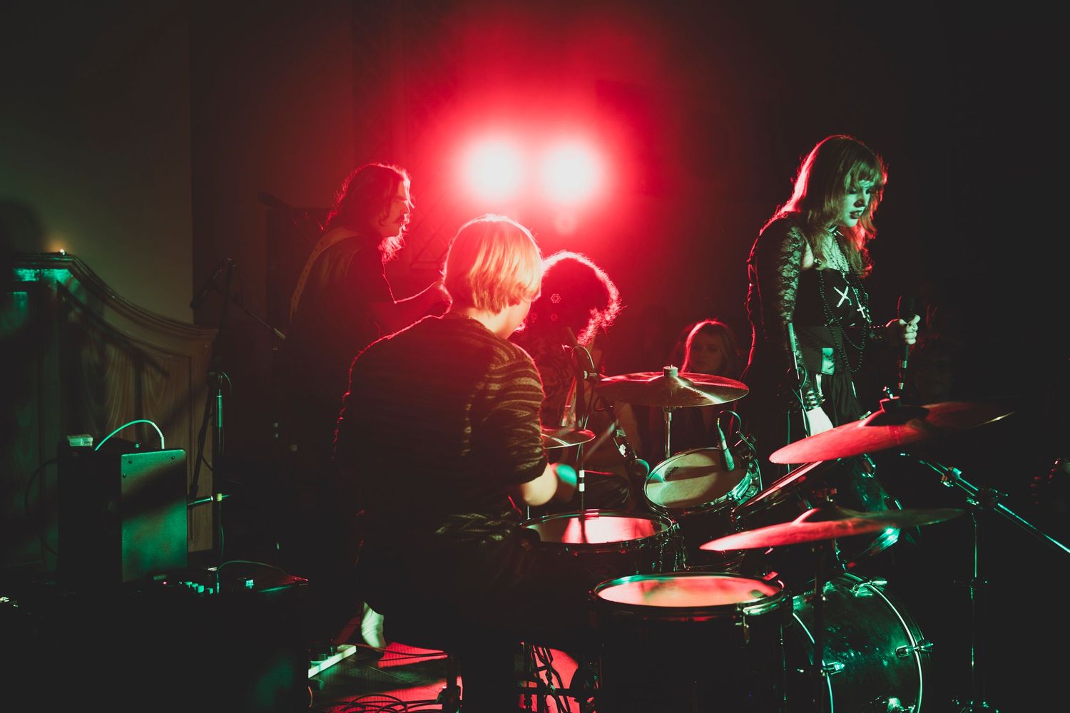 Band performing on stage with red lighting and silhouetted musicians playing drums.