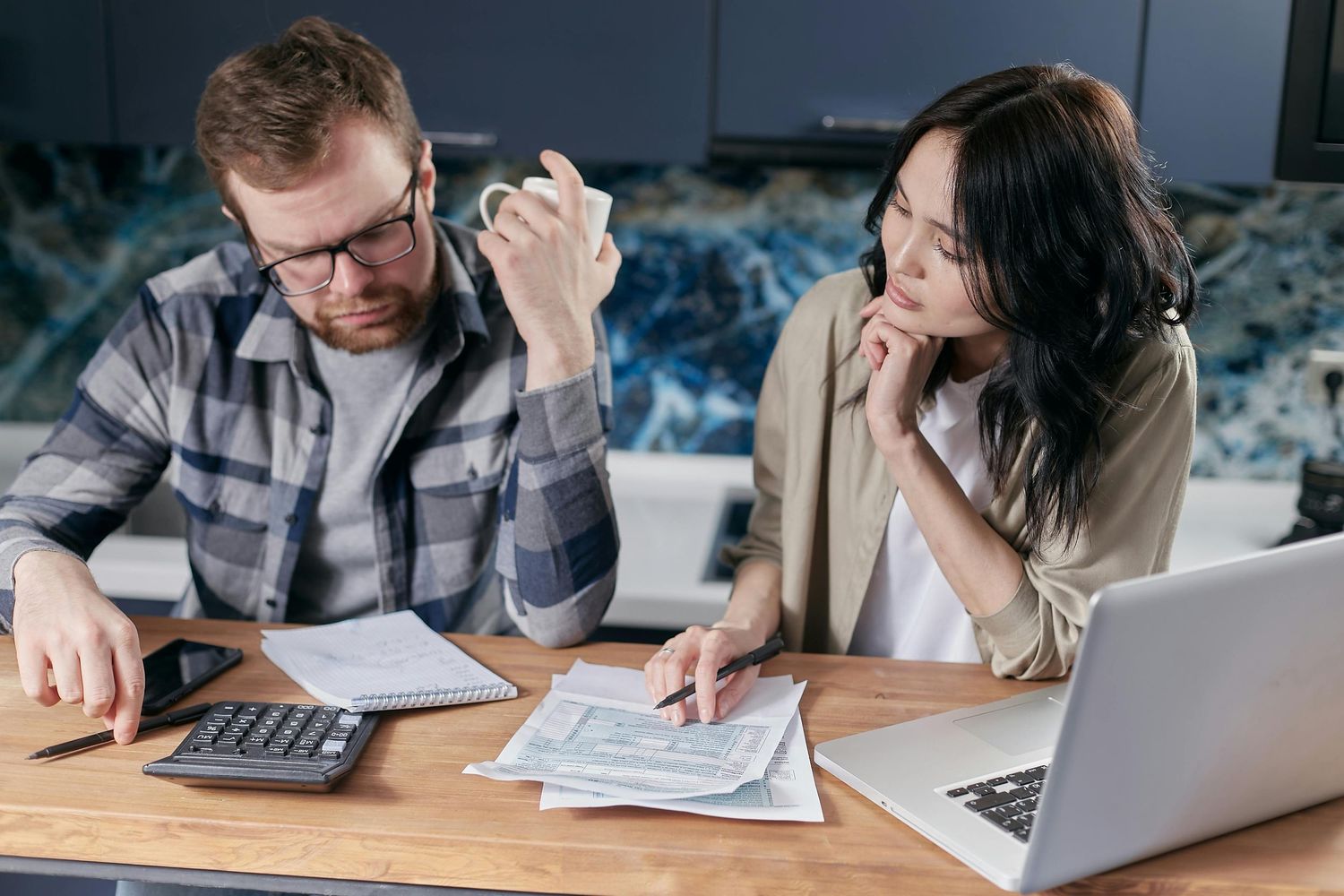 Two people review financial paperwork and documents together at a table with a calculator and laptop computer.