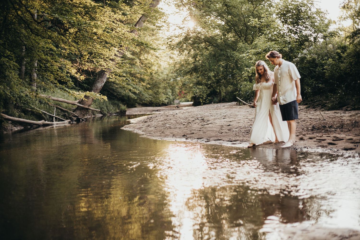 A couple embraces while standing in a serene creek surrounded by lush forest and golden sunlight.