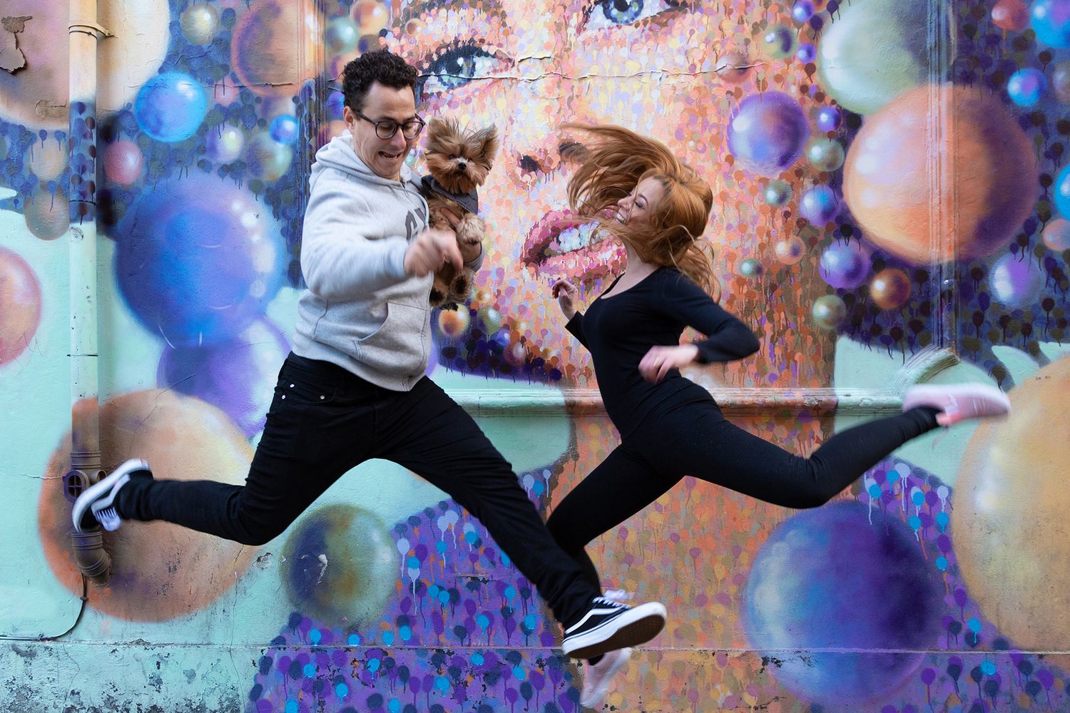 Two dancers leap joyfully in sync against a vibrant wall mural with colorful bokeh bubble effects.