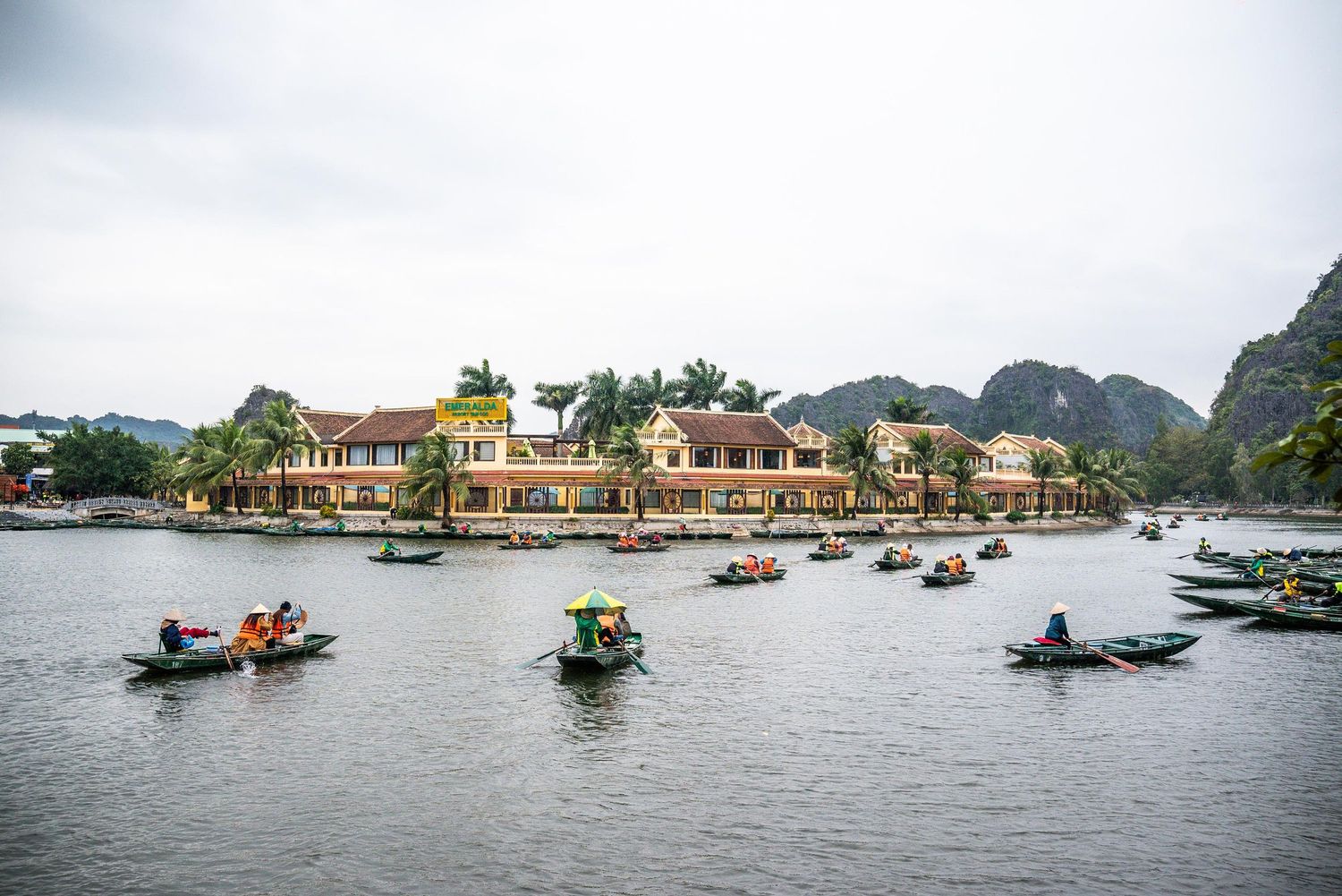 Small boats navigate the waterway near colorful buildings and limestone mountains in Tam Coc, Vietnam.
