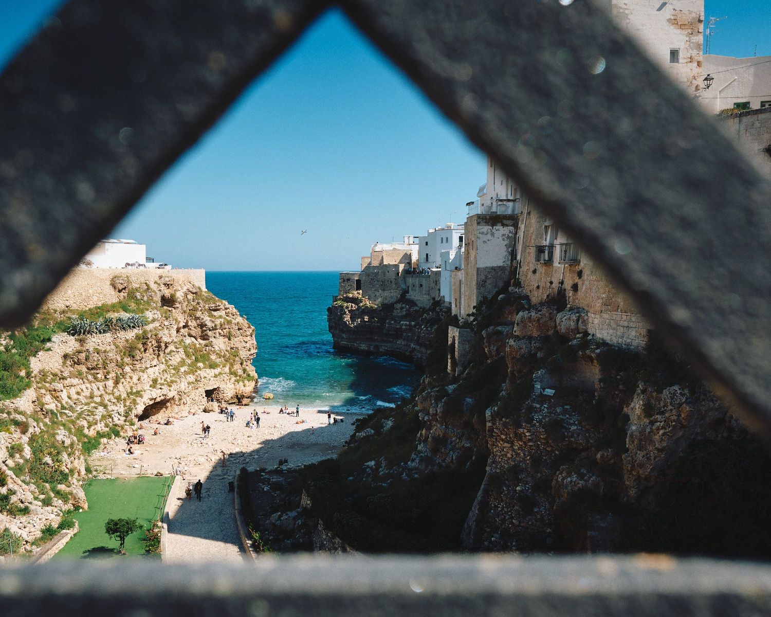 Scenic coastal view through metal railings showing rocky cliffs and turquoise Mediterranean waters against a clear blue sky.