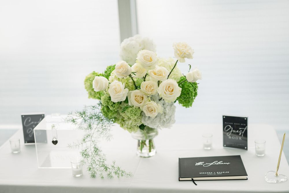 Elegant white rose and hydrangea centerpiece with green accents sits beside black guest books on white table.