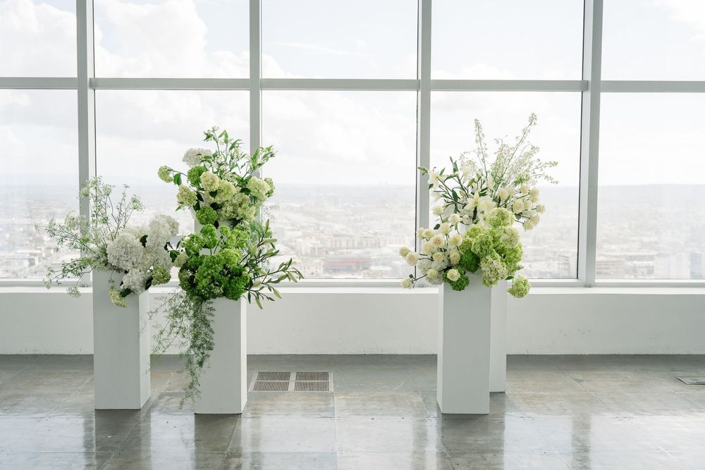 Three tall white floral arrangements in modern vases against a bright window with city views.