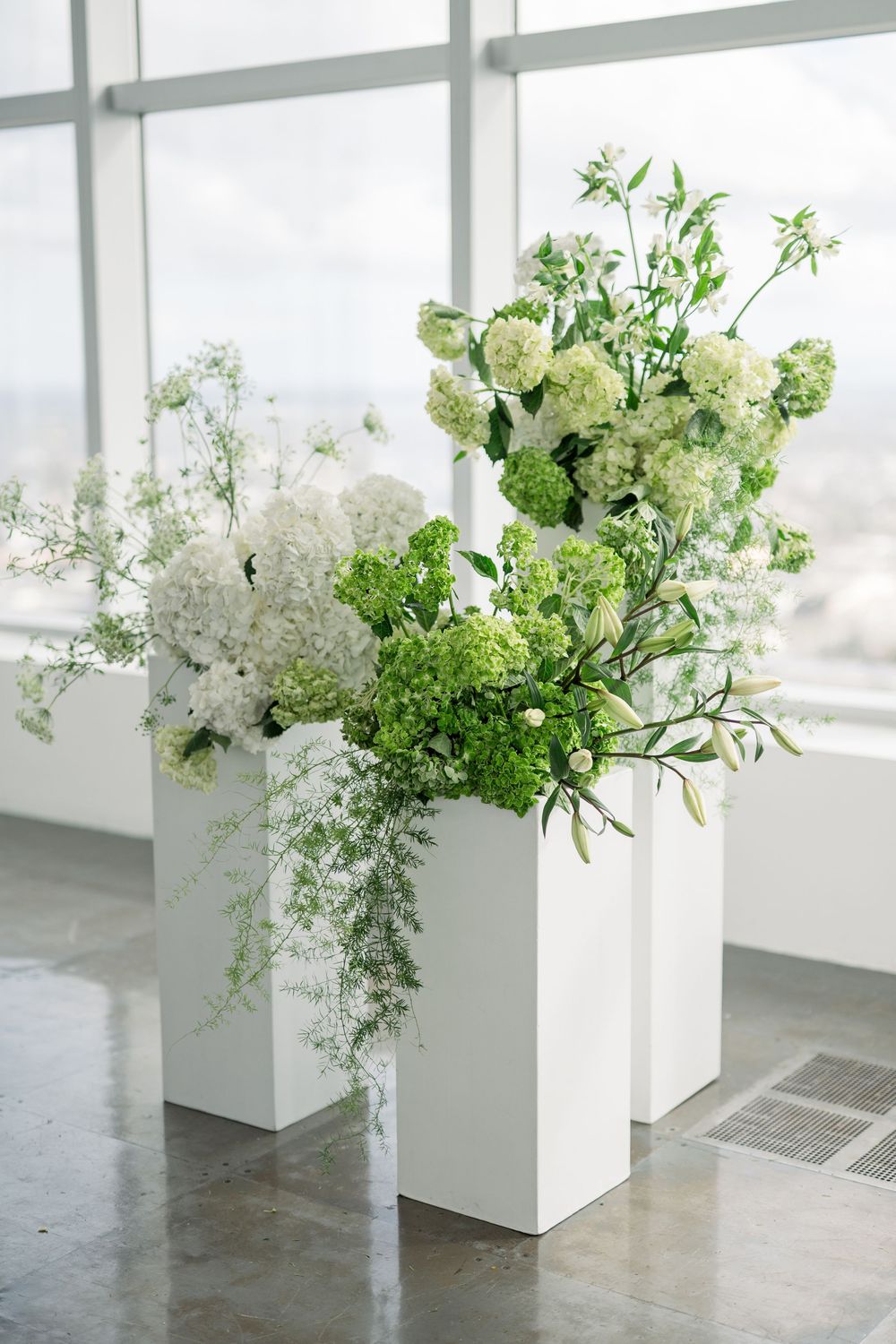 Modern white pedestal arrangements with green and white flowers against bright windows.