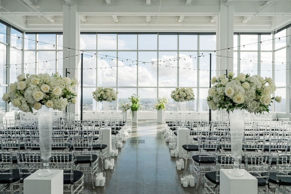Elegant white wedding ceremony setup with floral arrangements and ghost chairs in a bright venue.