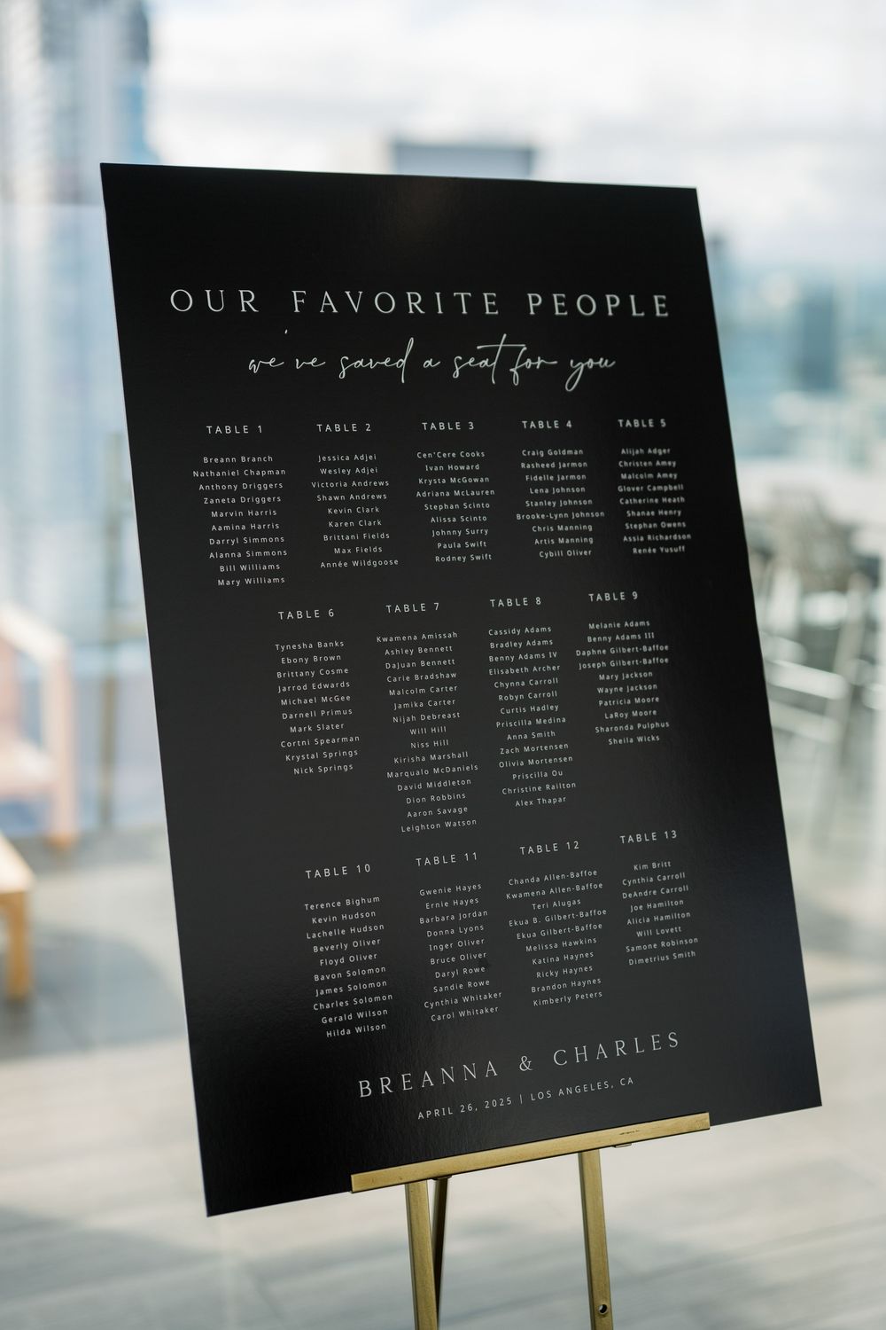 Modern black and white seating chart design displays guest table assignments on an easel with city views behind.