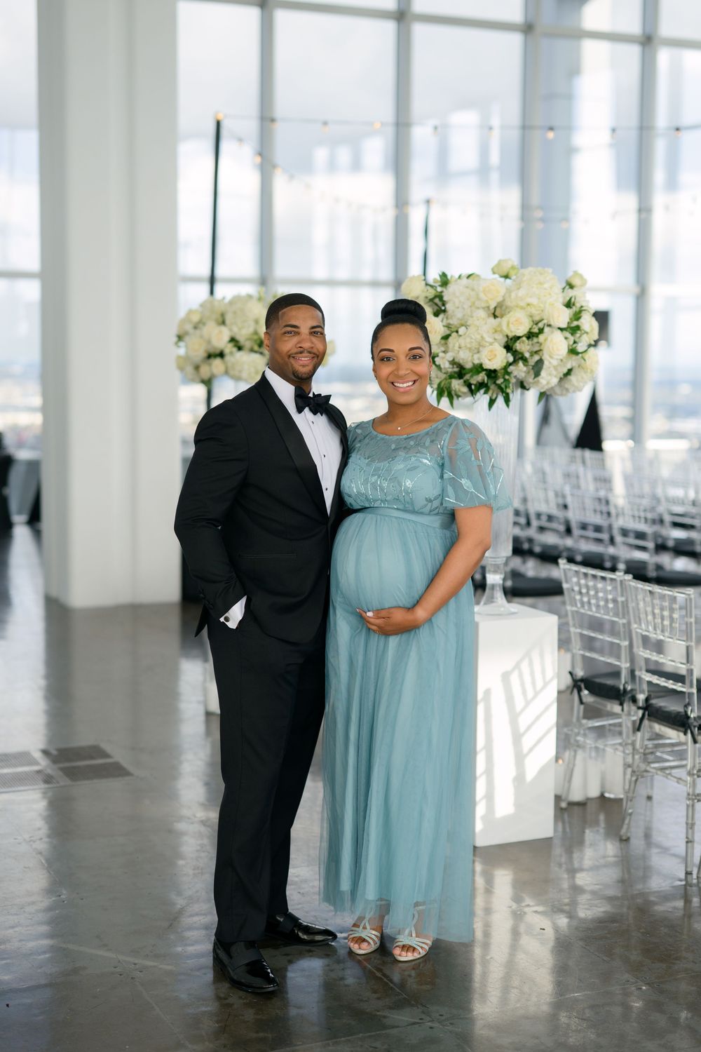 A couple poses in an elegant venue with white floral arrangements, the woman wearing a flowing light blue gown.