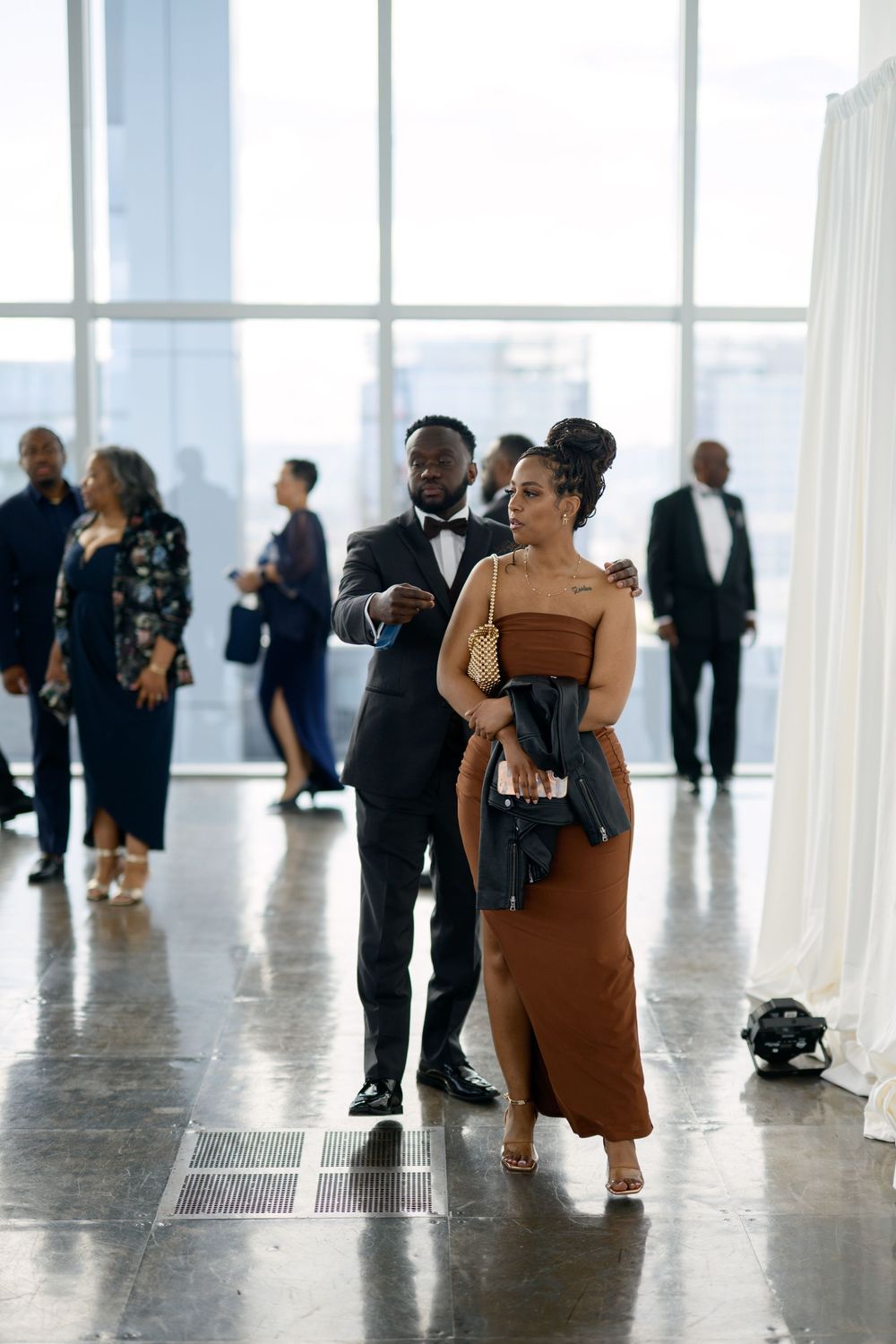 Wedding guests in formal attire mingle in a modern venue with floor-to-ceiling windows.