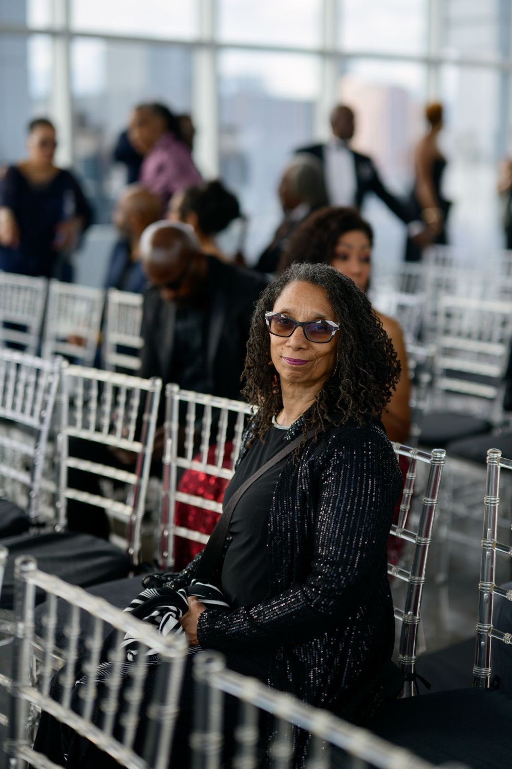 A stylish guest in a black outfit and glasses sits in white chairs at an elegant event space with large windows.
