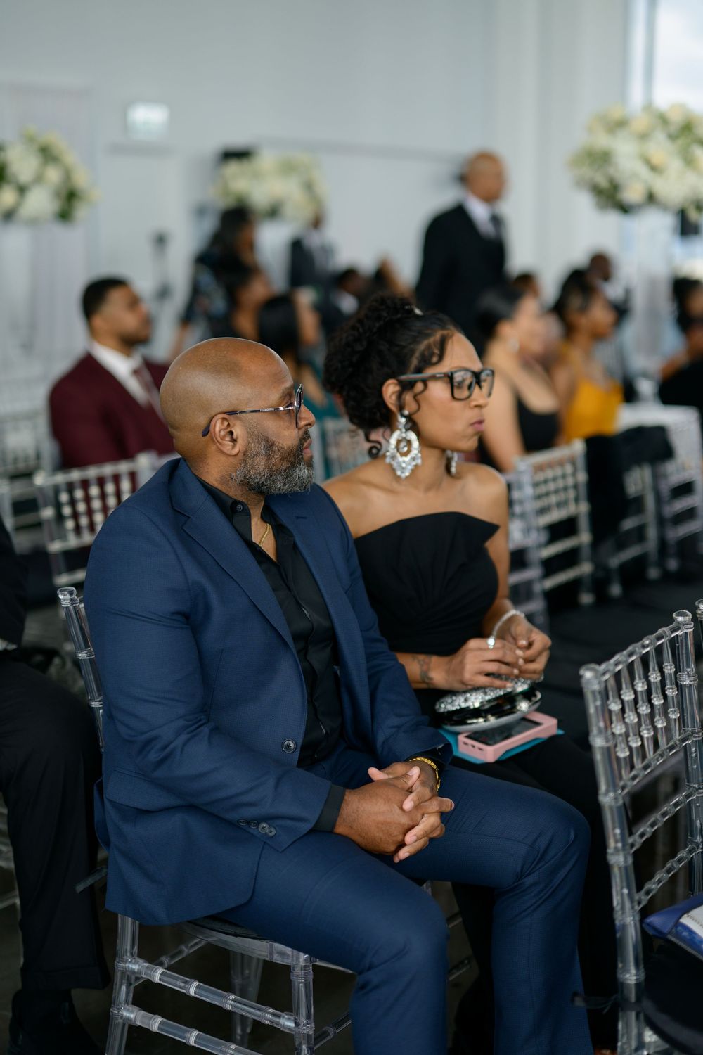 Well-dressed guests seated in silver chairs at an elegant formal event with white floral arrangements.