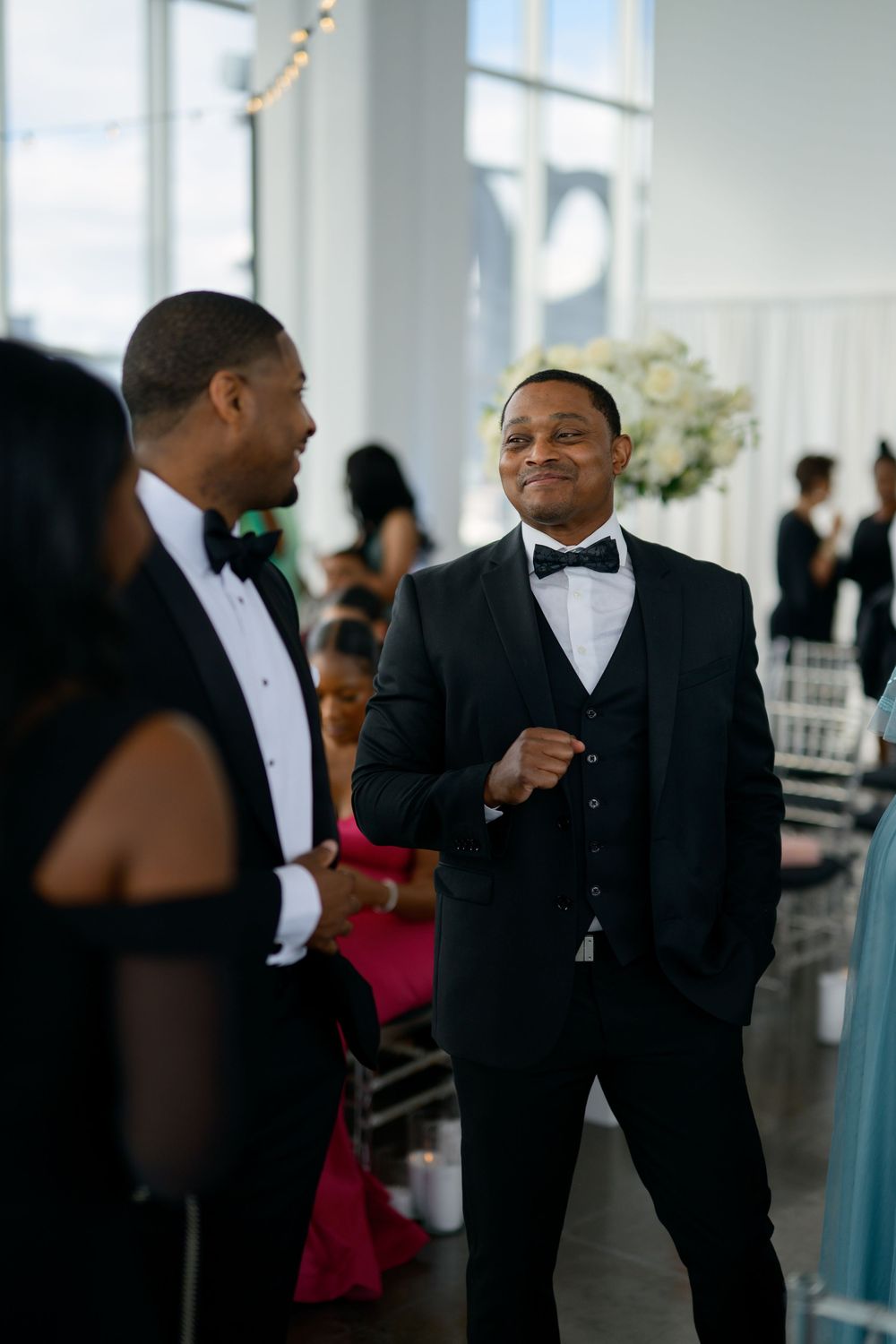 Two people in formal black tuxedos and bow ties engaged in conversation at an elegant event venue.