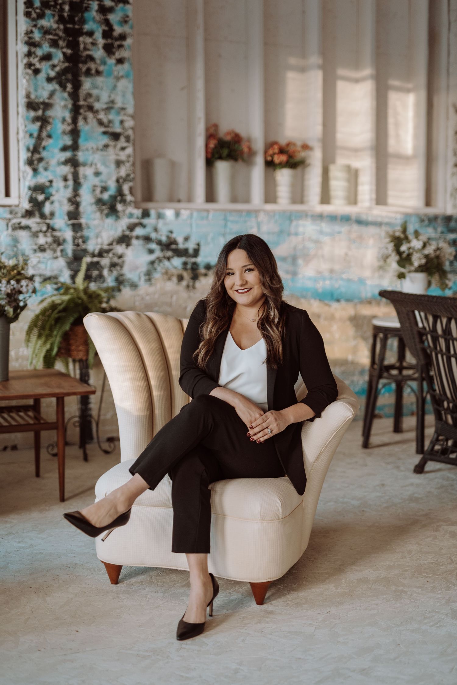 Professional business portrait of someone seated in a cream armchair wearing black suit and white top in an airy studio space.