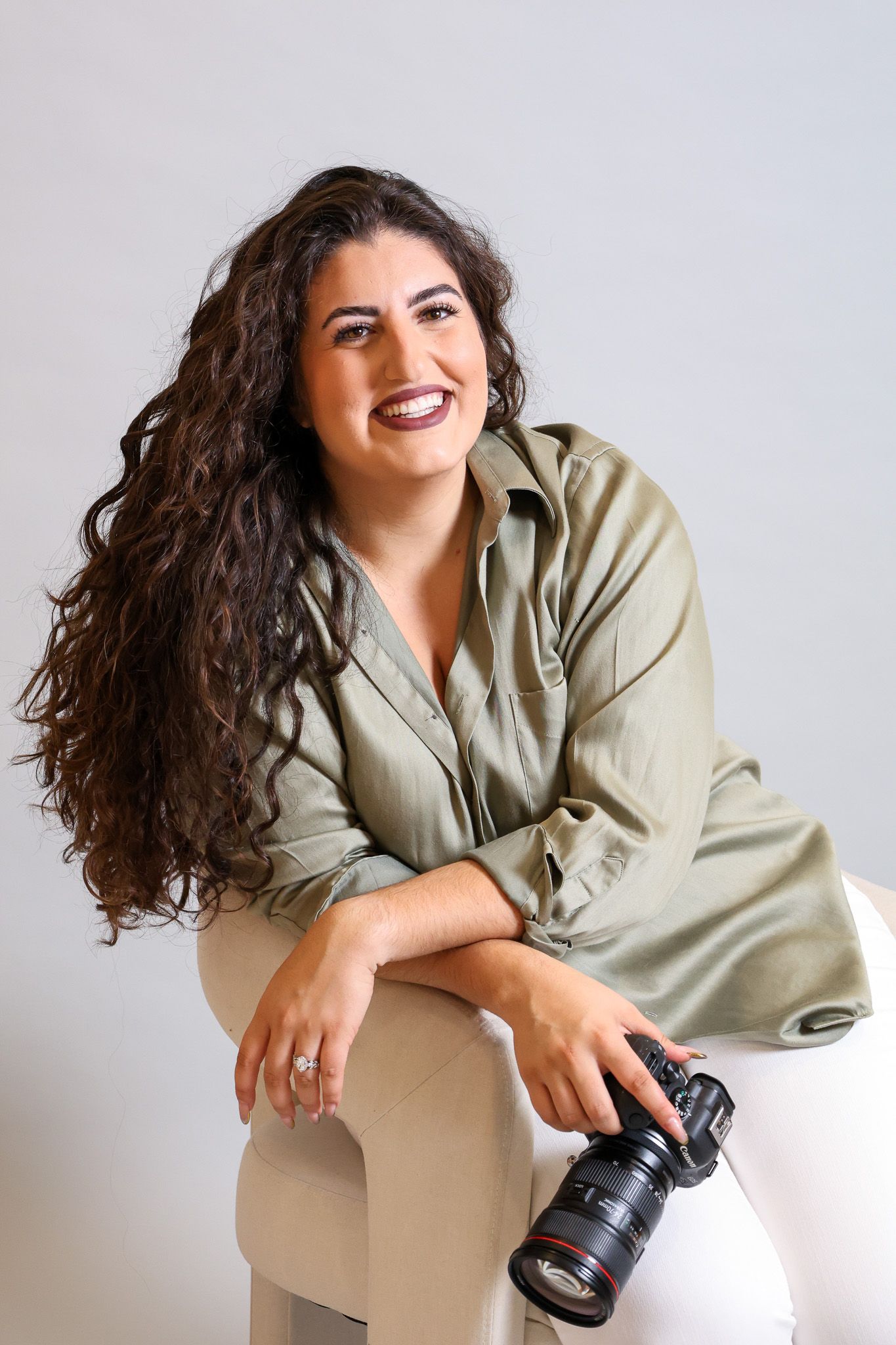 Professional photographer with long curly dark hair in beige blouse holding camera against white background.