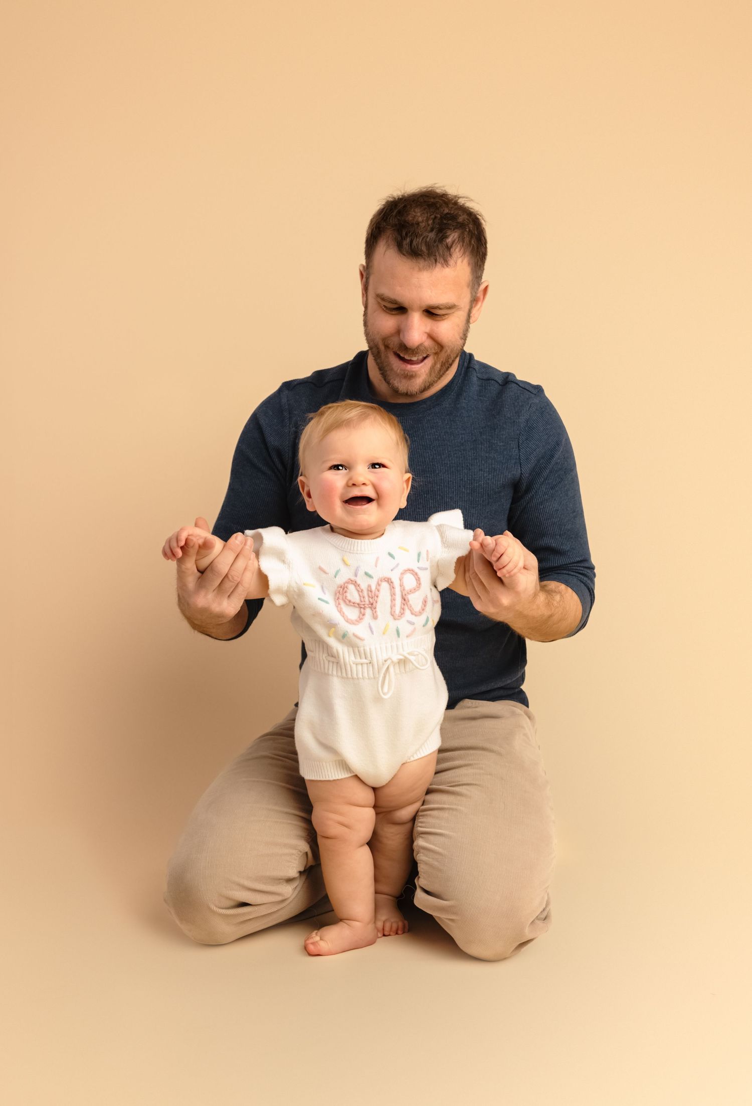 Dad and one year-old baby girl smiling against a cream background during a cake smash session in Charlottesville, Virginia.