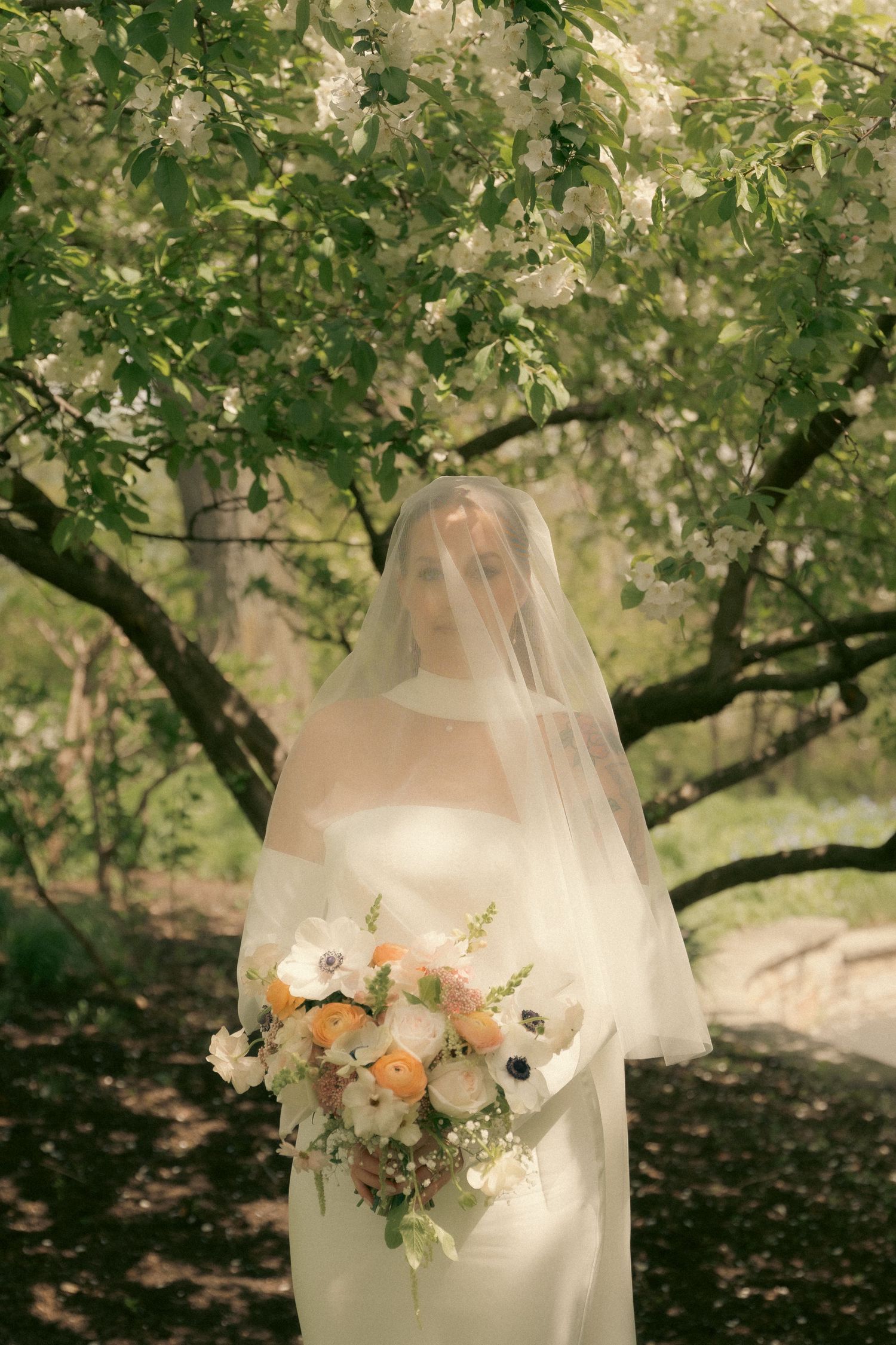 A romantic bridal figure in white dress holds a soft peach and white floral bouquet under flowering spring trees.