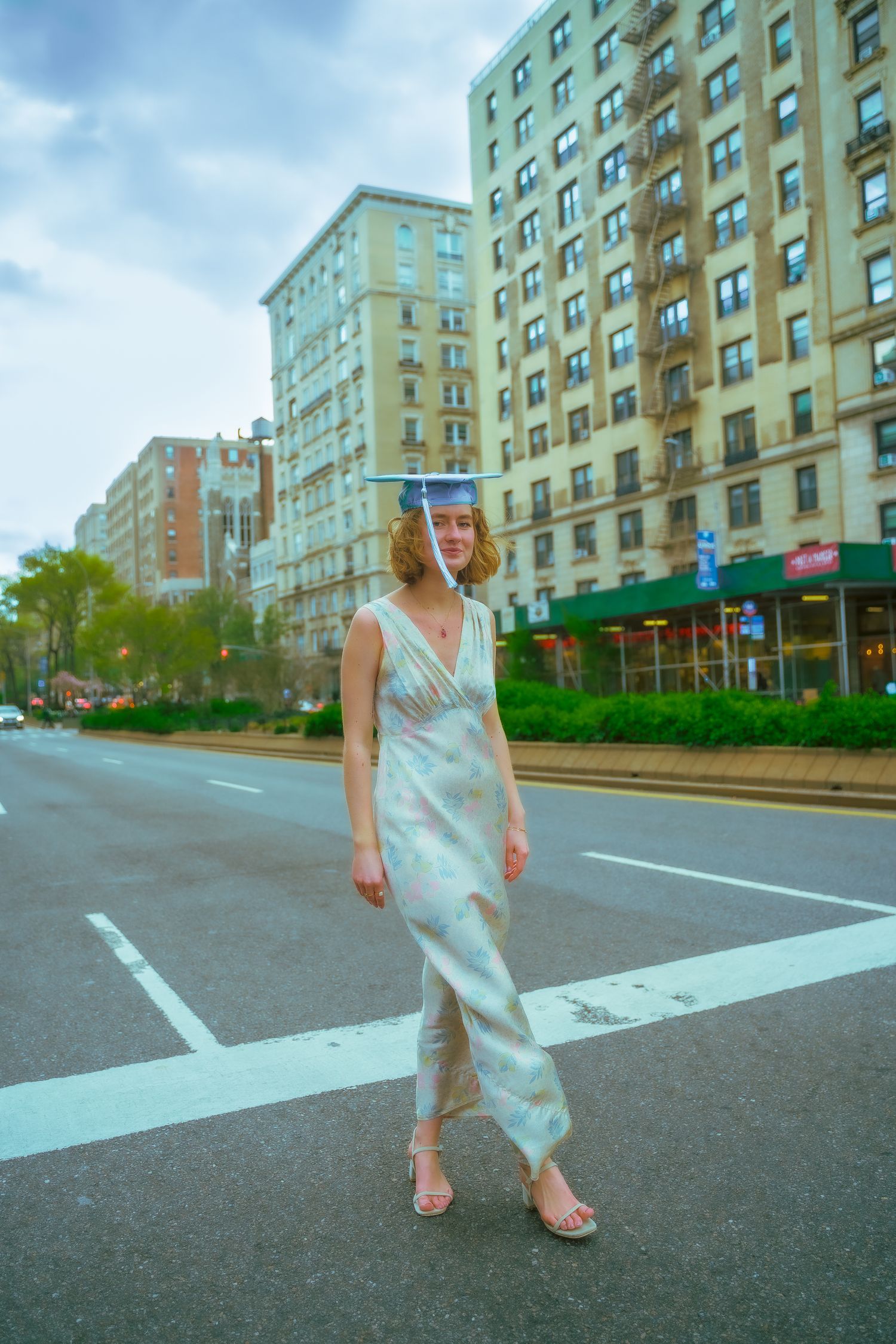 Graduate in silver dress poses on urban street with tall apartment buildings in background.