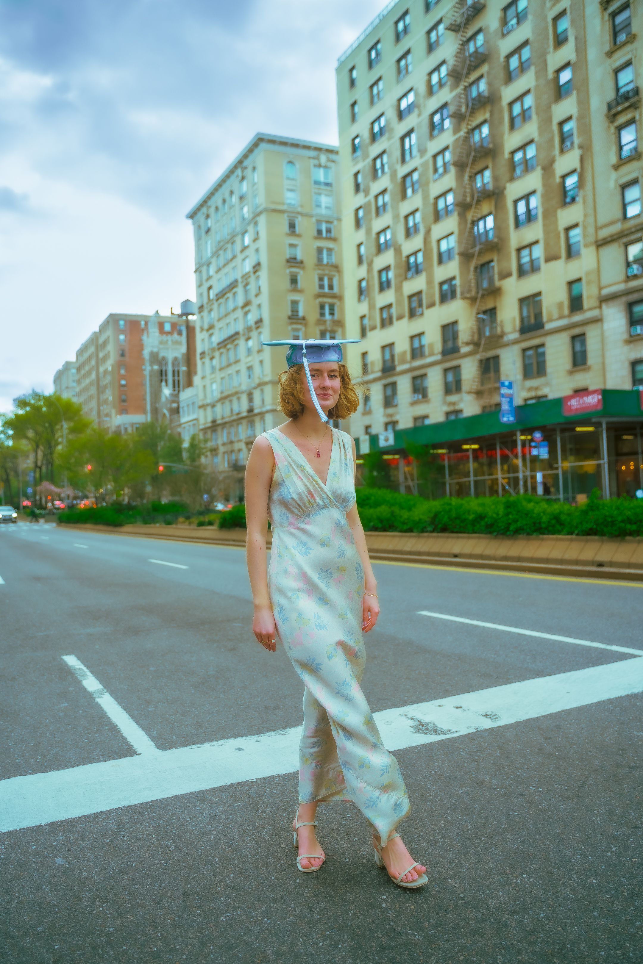 Graduate in silver dress poses on urban street with tall apartment buildings in background.