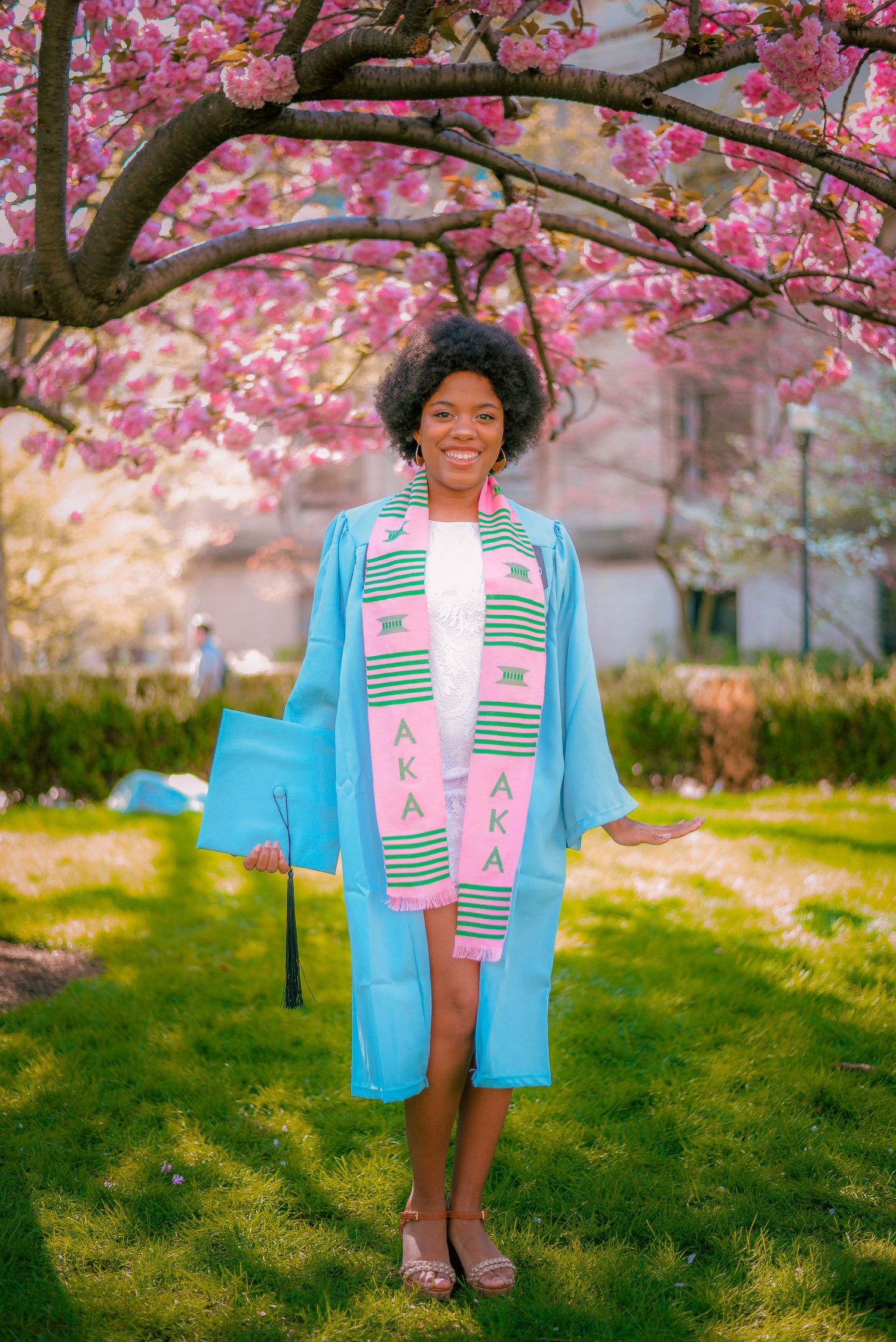 Graduate wearing patterned dress and light blue graduation gown stands beneath pink cherry blossoms.