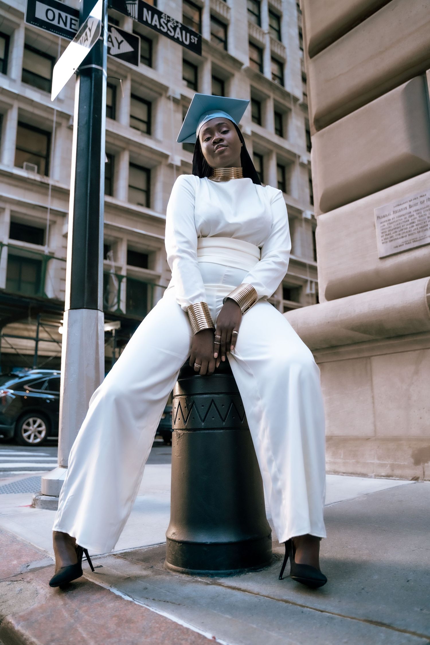 Person in all-white outfit sits stylishly on a street pole in an urban setting.