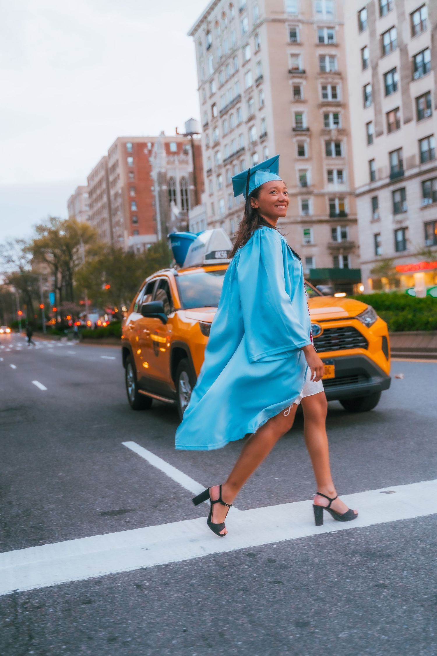 Graduate in light blue cap and gown twirls joyfully on street with yellow taxi in background.