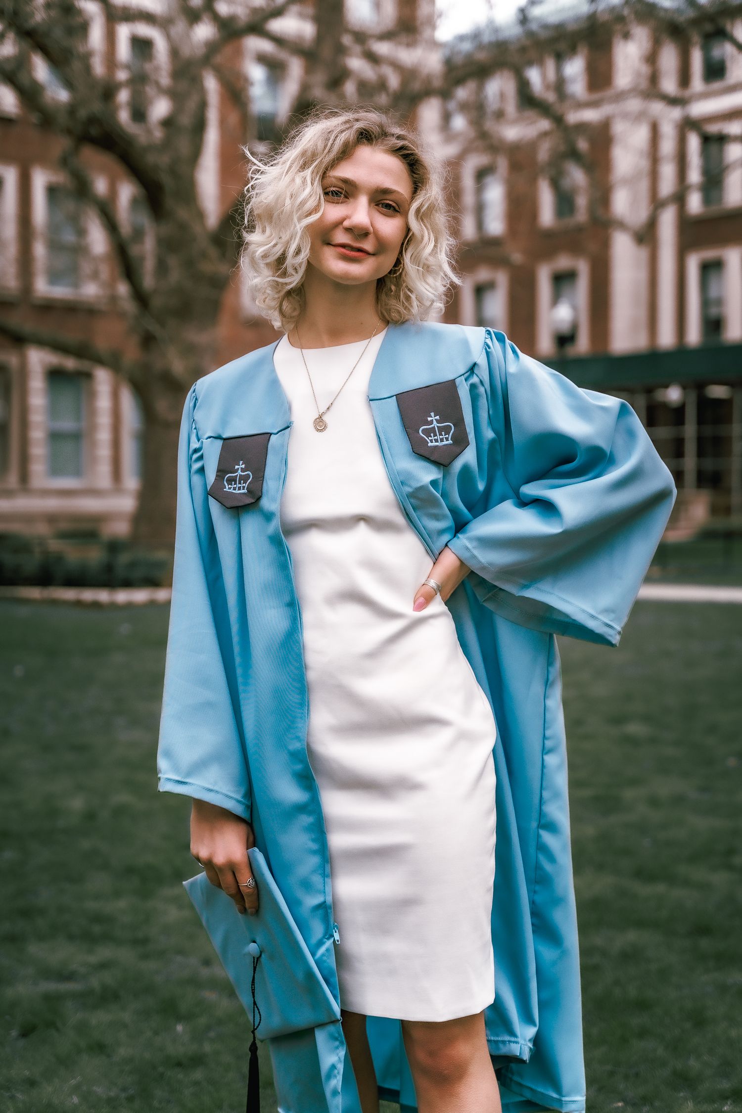 Graduate in light blue gown and white dress poses confidently on campus grounds.