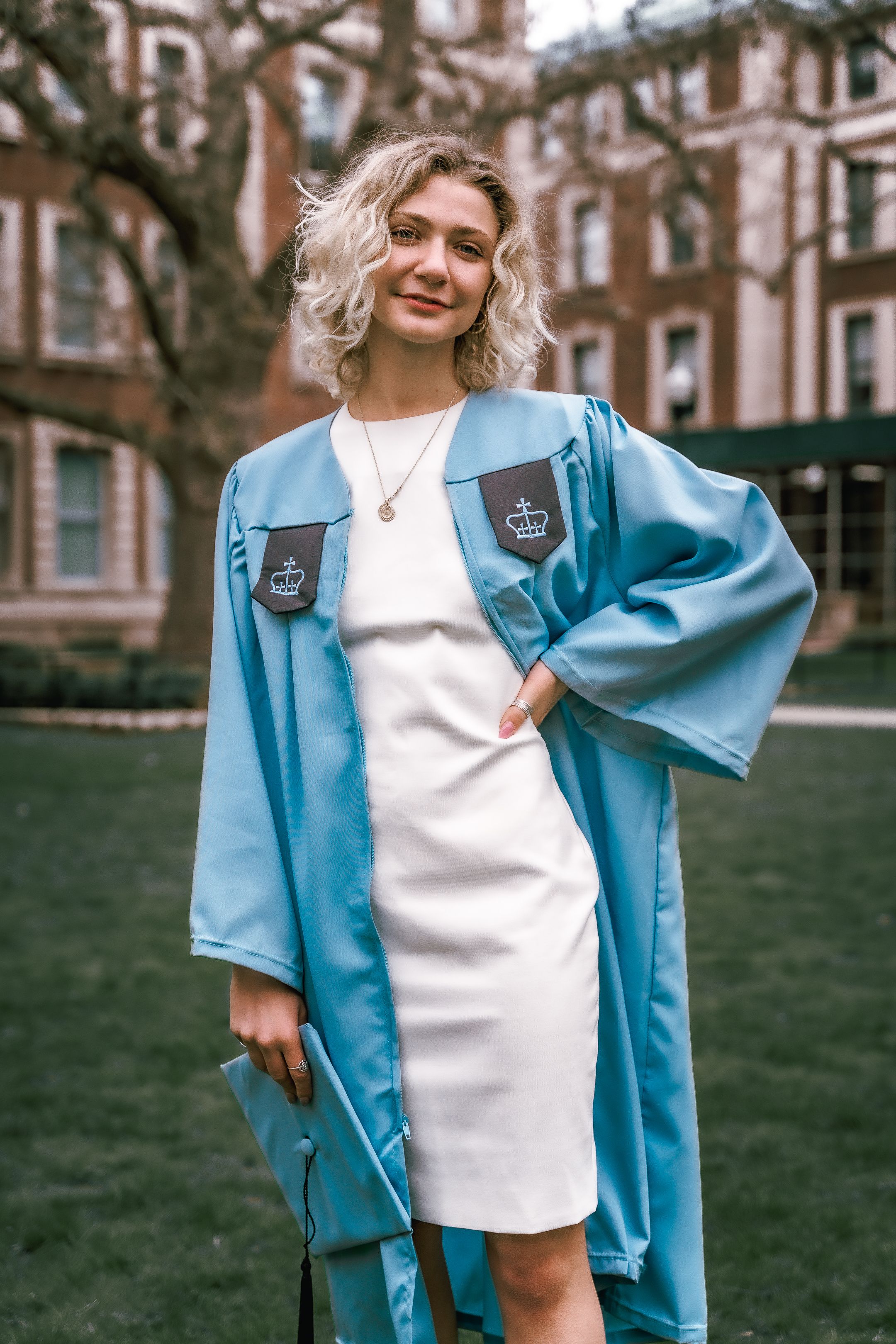 Graduate in light blue gown and white dress poses confidently on campus grounds.